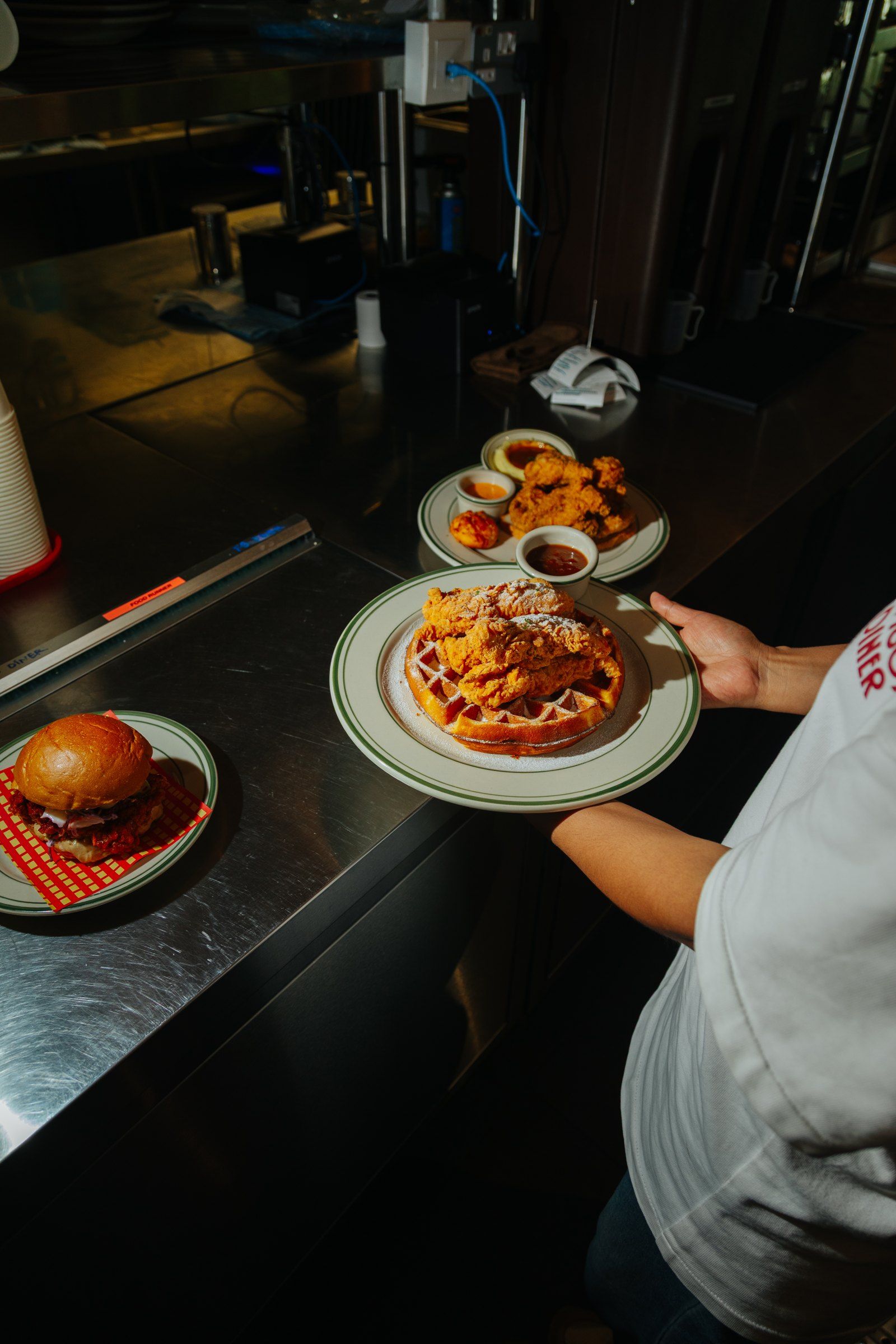 A server carries a chicken and waffle plate along the steel pass, a burger and a plate of tenders waiting beside it