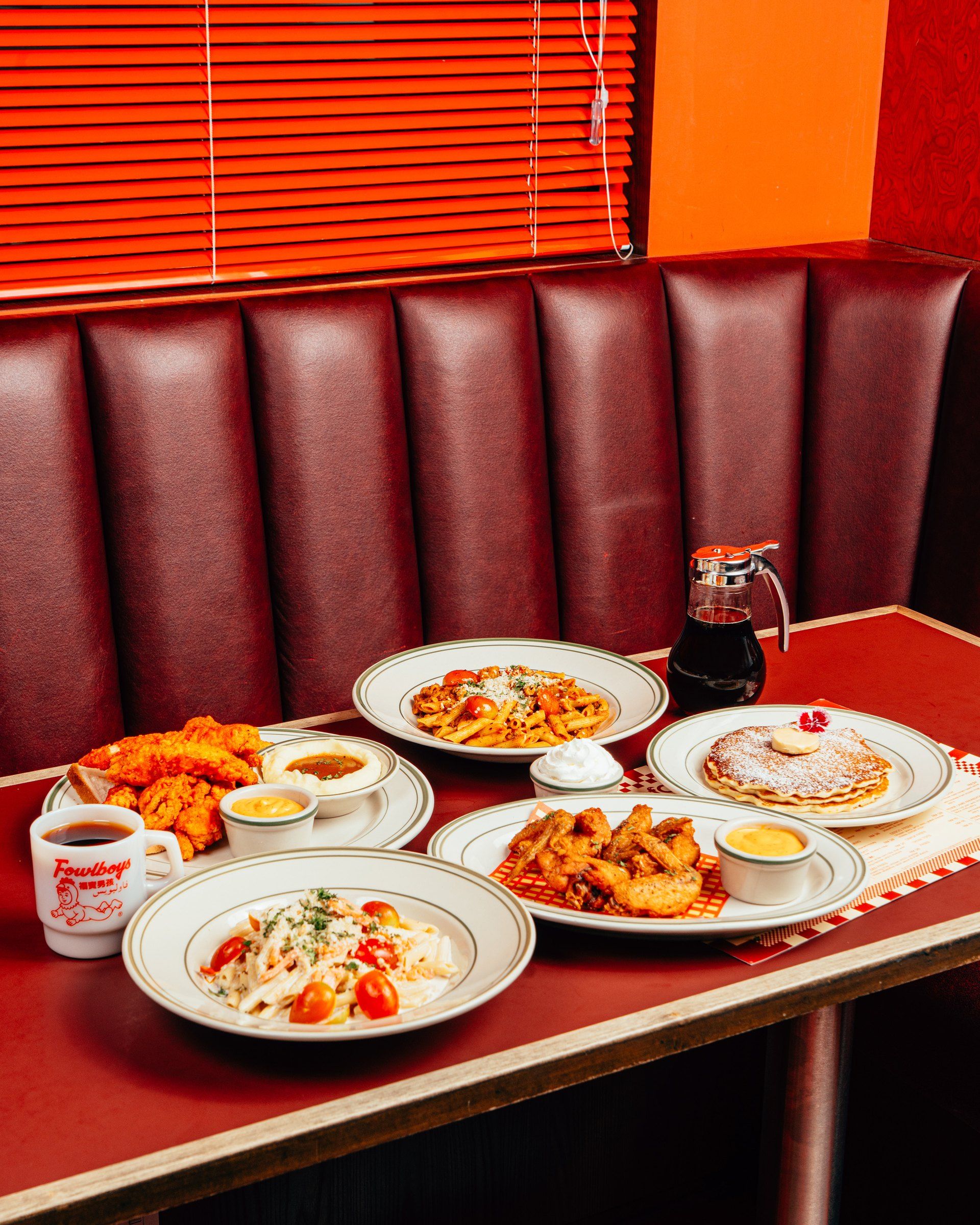 A booth by the orange blinds laid with tenders, pastas, wings, pancakes, mash and coffee